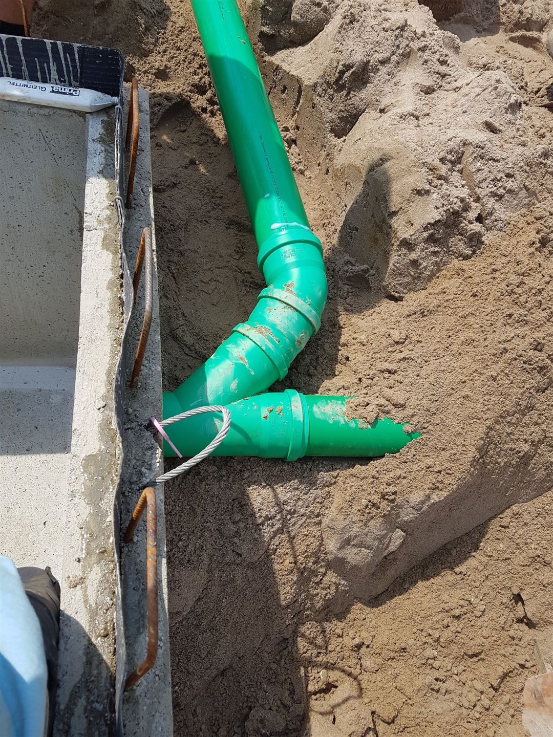 Grüne Abwasserleitung im Sand neben einer Kellerwand mit schwarzer Abdichtung und Schutzlage.