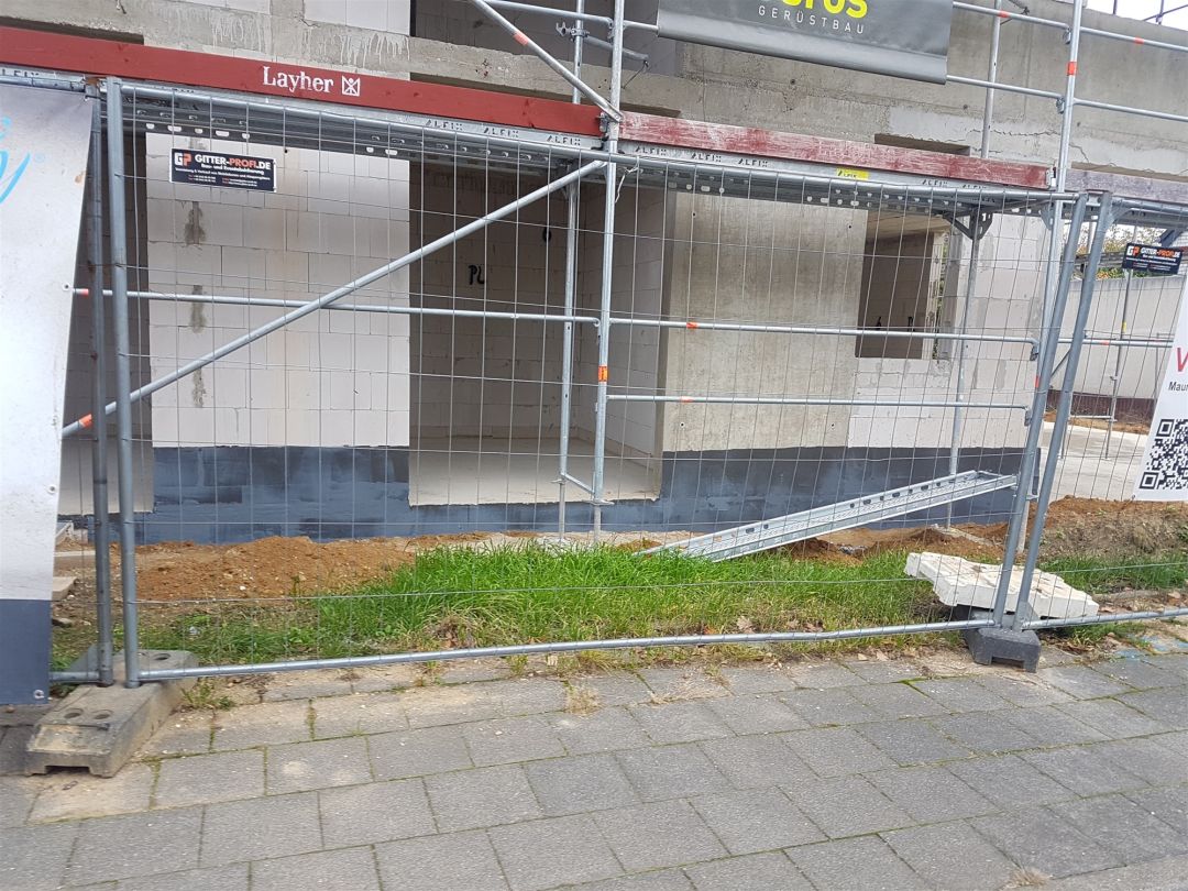 Gerüst vor der Fassade eines Gebäudes mit ausgesparter Wandöffnung, davor ein gepflasterter Gehweg.