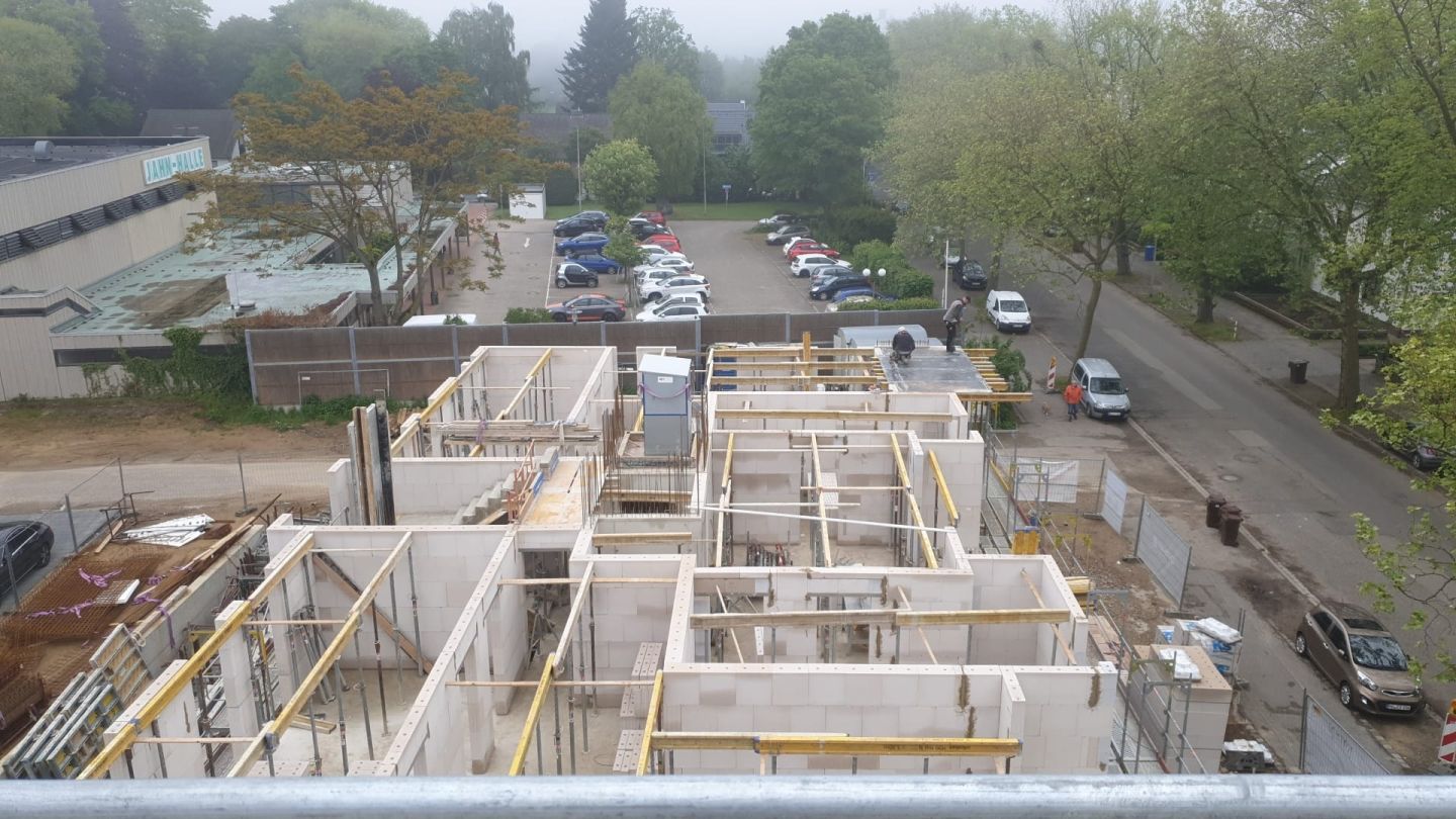 Blick von oben auf einen Rohbau mit Kellerwänden, dahinter eine Wohnstraße mit parkenden Autos und Bäumen.